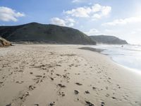 a photo taken on a sandy beach with footprints left in the sand and hills in the background