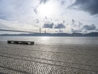 the bench is empty near the water under a large bridge and a cloudy sky with dark clouds