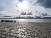 the bench is empty near the water under a large bridge and a cloudy sky with dark clouds