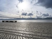 the bench is empty near the water under a large bridge and a cloudy sky with dark clouds