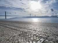 an old stone floor in front of water and a bridge, with sun shining in the sky