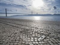 an old stone floor in front of water and a bridge, with sun shining in the sky