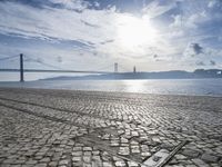 an old stone floor in front of water and a bridge, with sun shining in the sky