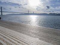 a woman is walking along the beach near the water and the bridge that is under a blue sky