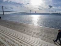 a woman is walking along the beach near the water and the bridge that is under a blue sky