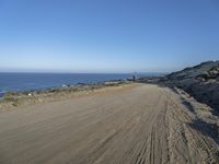 a dirt road beside a shore line with a distant ocean in the background on a sunny day