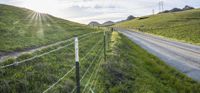 a road and fence with a grassy hill in the distance with a field and sun in the distance