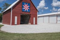a barn on top of a gravel road next to grass and trees in front of a blue sky