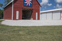 a barn on top of a gravel road next to grass and trees in front of a blue sky