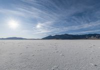 the view of a snow covered field with footprints in the dirt and a sun beaming behind the mountains