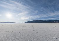 the view of a snow covered field with footprints in the dirt and a sun beaming behind the mountains