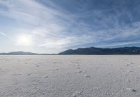 the view of a snow covered field with footprints in the dirt and a sun beaming behind the mountains