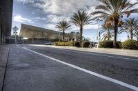 an empty asphalt road surrounded by palm trees and building in the background with a cloudy sky