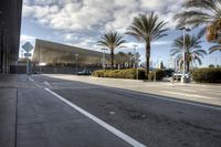 an empty asphalt road surrounded by palm trees and building in the background with a cloudy sky