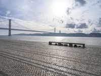 stone pavement beside the water and under a bridge with bridge in the distance and clouds in the sky