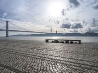 stone pavement beside the water and under a bridge with bridge in the distance and clouds in the sky