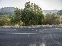 an asphalt road runs next to green trees and hill side with a mountain range in the background