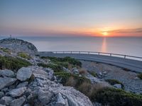 a curved road going along the sea as the sun set on the horizon behind it