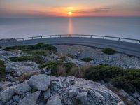 a curved road going along the sea as the sun set on the horizon behind it