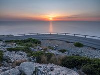 a curved road going along the sea as the sun set on the horizon behind it