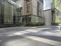 two people walk on a sunny street near an architectural structure with glass walls and stairs