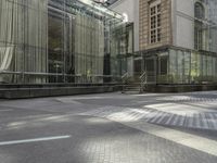 two people walk on a sunny street near an architectural structure with glass walls and stairs