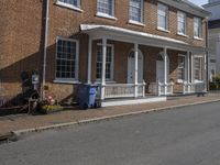 a brick house with white windows and windows next to a sidewalk with a blue trash can on one side of the street