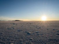 the sun shines brightly behind a frozen landscape with footprints in the snow and hills in the distance