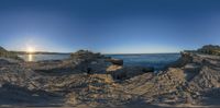 fish - eye photo of a large ocean with some rocks at the beach and the sun rising behind it