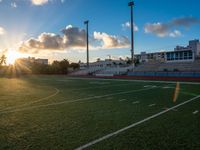 an empty field with a football stadium with seats on each side and the sun setting behind it