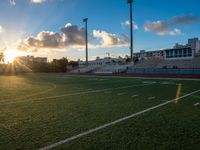 an empty field with a football stadium with seats on each side and the sun setting behind it