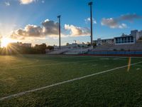 an empty field with a football stadium with seats on each side and the sun setting behind it