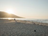 the sun is rising over the water, and the people on the beach are walking toward the shore