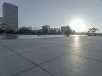 a fountain in the middle of a public park at sunrise time with the sun setting over buildings