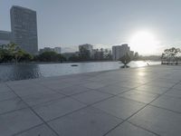 a fountain in the middle of a public park at sunrise time with the sun setting over buildings