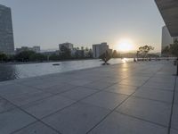 a fountain in the middle of a public park at sunrise time with the sun setting over buildings