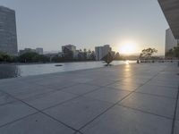 a fountain in the middle of a public park at sunrise time with the sun setting over buildings