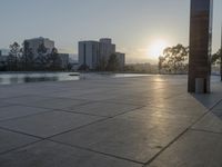 a fountain in the middle of a public park at sunrise time with the sun setting over buildings