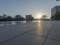 a fountain in the middle of a public park at sunrise time with the sun setting over buildings