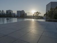 a fountain in the middle of a public park at sunrise time with the sun setting over buildings