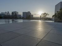 a fountain in the middle of a public park at sunrise time with the sun setting over buildings