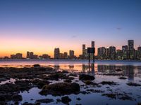 an ocean shore is lined with rocks and some lights as the sun rises over the city