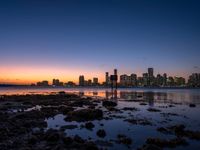 an ocean shore is lined with rocks and some lights as the sun rises over the city