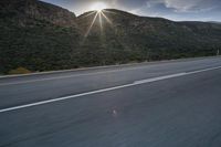 the sun is rising on a road surrounded by mountains and trees on the side of the road