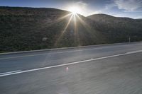 the sun is rising on a road surrounded by mountains and trees on the side of the road