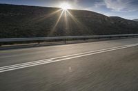 the sun is rising on a road surrounded by mountains and trees on the side of the road