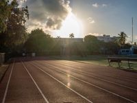 a sunrise at a track and field with some benches near it and a fence with the sun rising in the background