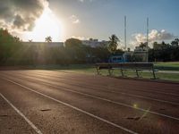 a sunrise at a track and field with some benches near it and a fence with the sun rising in the background