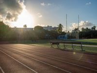 a sunrise at a track and field with some benches near it and a fence with the sun rising in the background