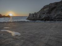 a lone motorcycle is parked near a rock formation on the beach at sunset, with water and ocean in the background